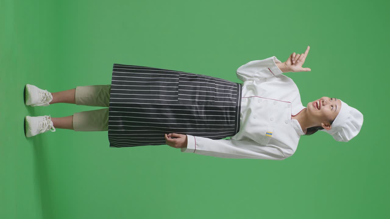 Full Body Of Asian Woman Chef Smiling And Pointing To Side While Standing In The Green Screen Background Studio