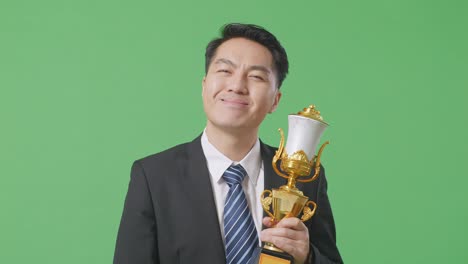 close up of asian business man in a suit and tie with a gold medal looking at a gold trophy in his hands and smiling to camera as the first winner on green screen background in the studio