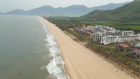 el hotel fantasma de lang co se sienta ocioso mientras las olas se estrellan en la playa de arena estéril. drone retrocede 60 fps.