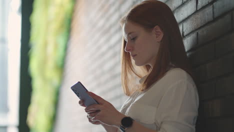 beautiful young business woman using phone in office. woman using a smartphone and leaning on a window texting sending emails planning meetings networking online browsing messages on mobile phone