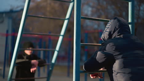 close-up of two people resting their hands on an iron bar, both wearing a hooded jacket and the other in the background blurred, with a blurred building in the distance