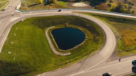 Luftdrohnenaufnahme-Einer-Ein--Und-Ausfahrtsrampe-Einer-Autobahnüberführung-Mit-Einem-Kleinen-Teich-In-Der-Mitte,-An-Dem-An-Einem-Sonnigen-Tag-Autos-Auf-Der-Autobahn-Fahren