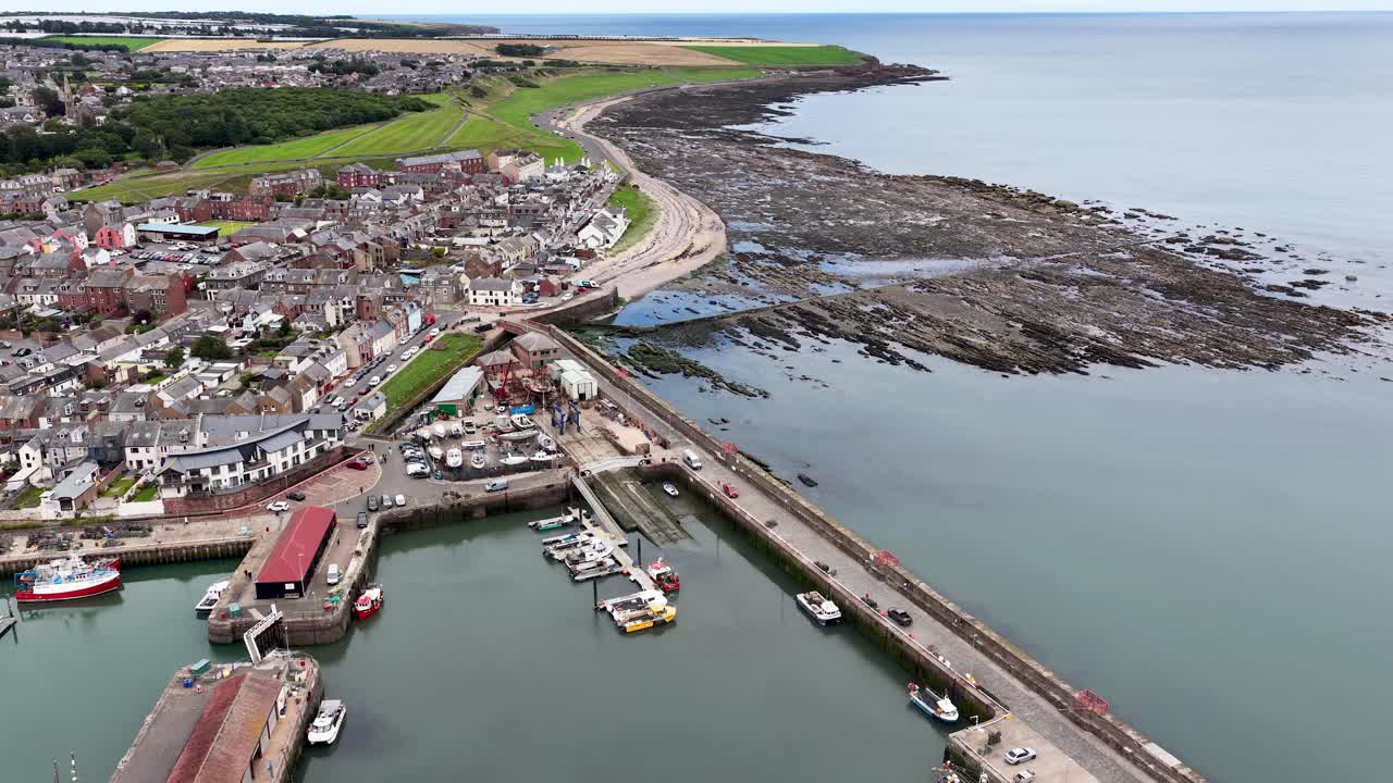 Drone camera glides above harbor, boats, and shoreline in a bright, overcast coastal town