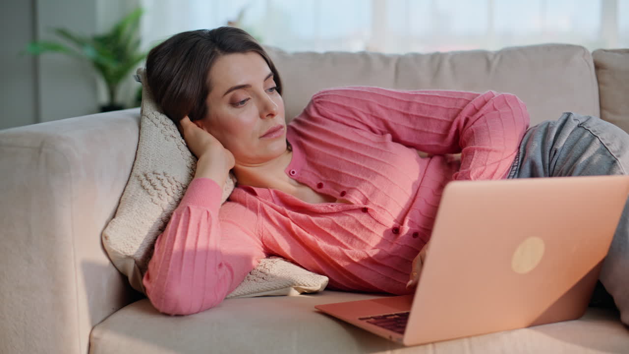 Bored woman browsing laptop lying comfortably at couch light living room closeup