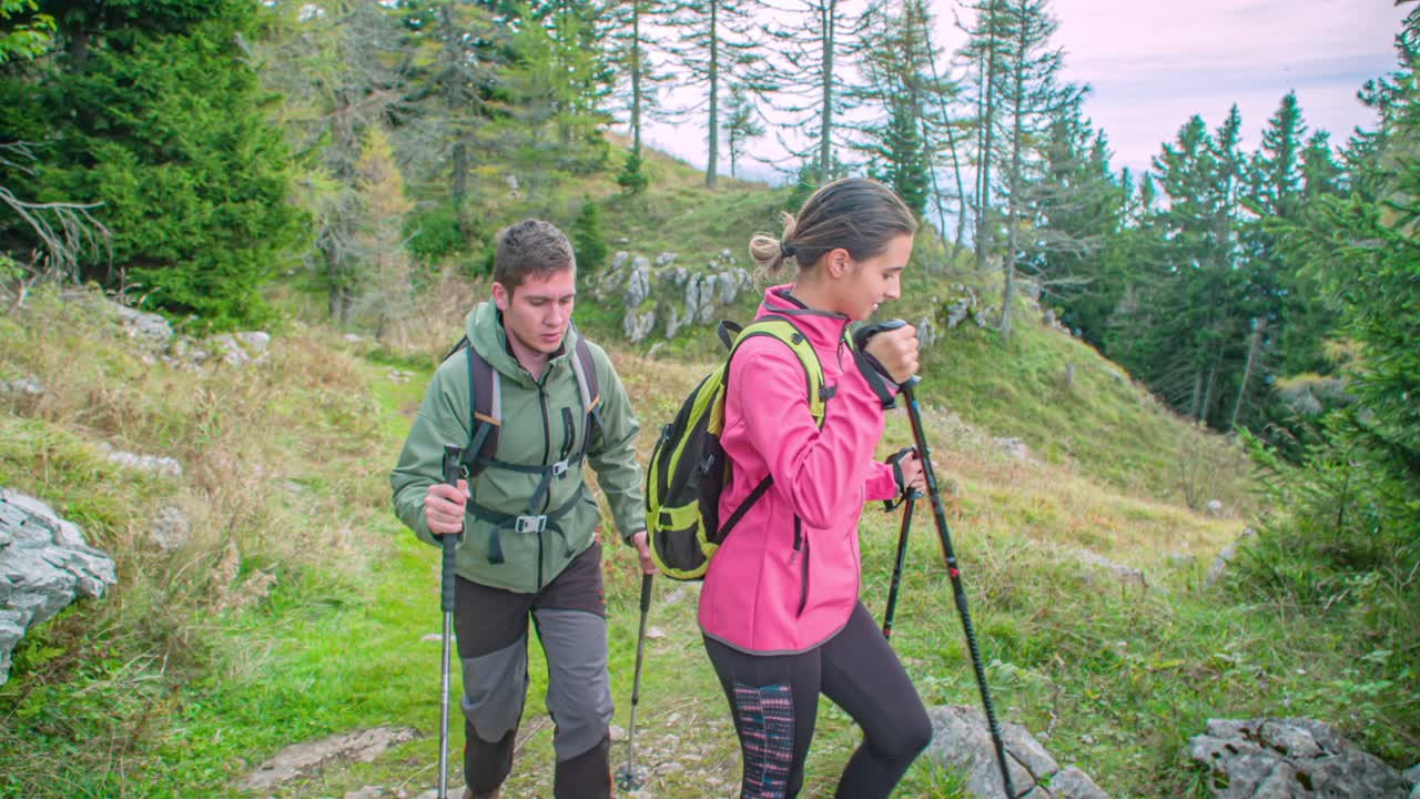 Young sporty couple hikes down rocky path with trekking sticks, stopping at information board. Trees in background