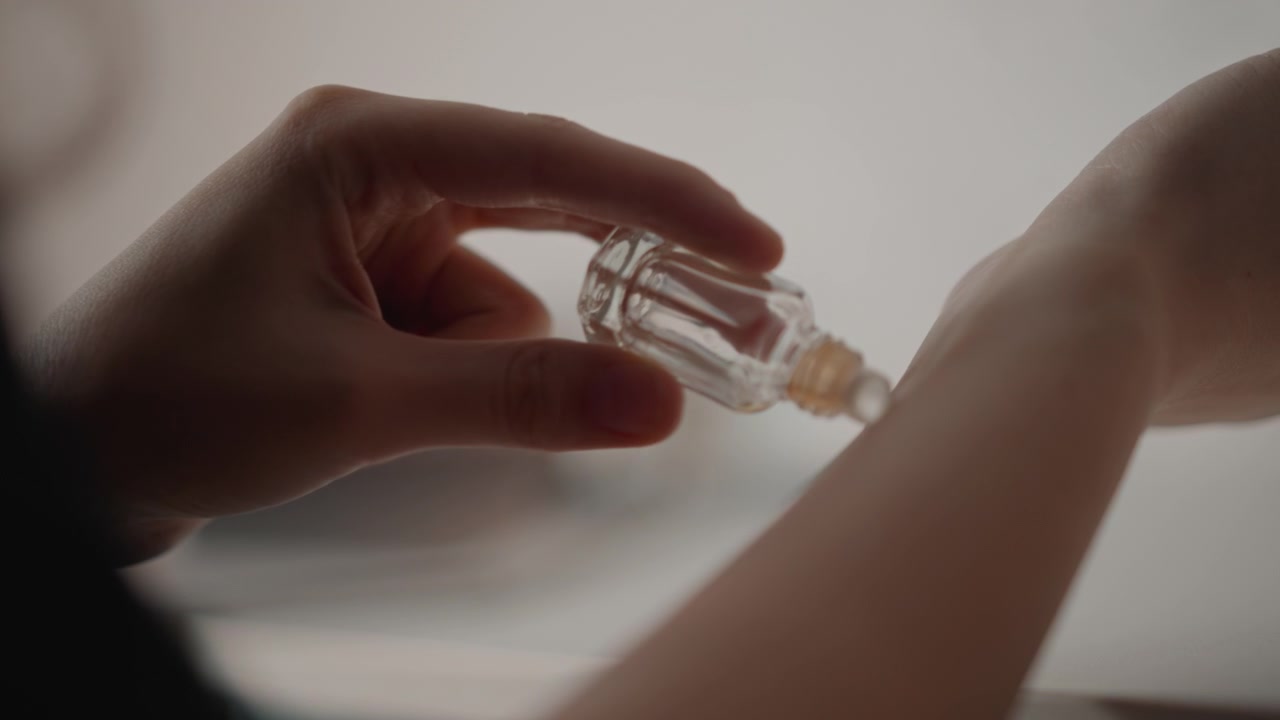 Inquadratura ravvicinata da sopra la spalla che mostra le mani che tengono una boccetta roll-on di vetro trasparente mentre la punta del roll-on tocca l'interno del polso e trasferisce una piccola goccia d'olio; le azioni visibili includono l'inclinazione della boccetta e la presa delle dita