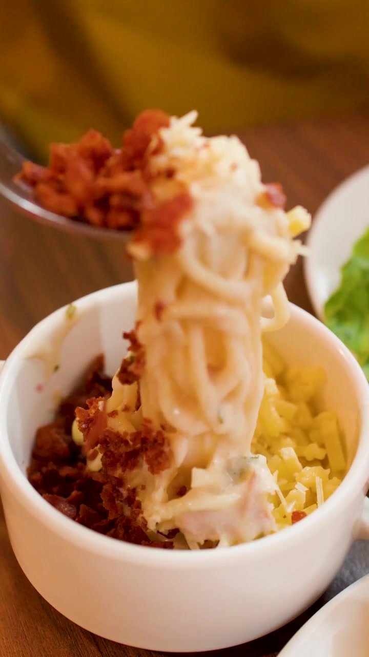 Primo piano di una forchetta che solleva pasta cremosa condita con pezzetti di pancetta e formaggio grattugiato.