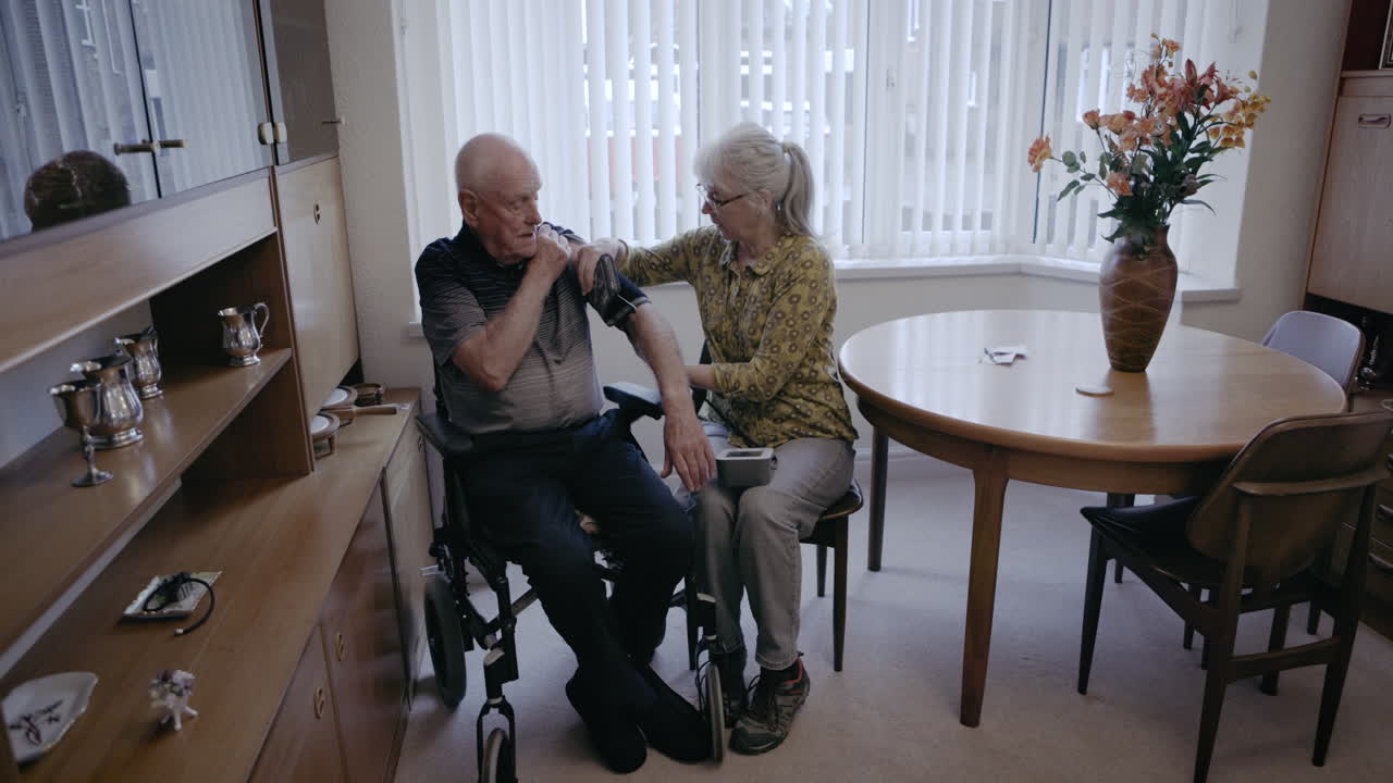 Elderly woman taking blood pressure of senior man in wheelchair at home