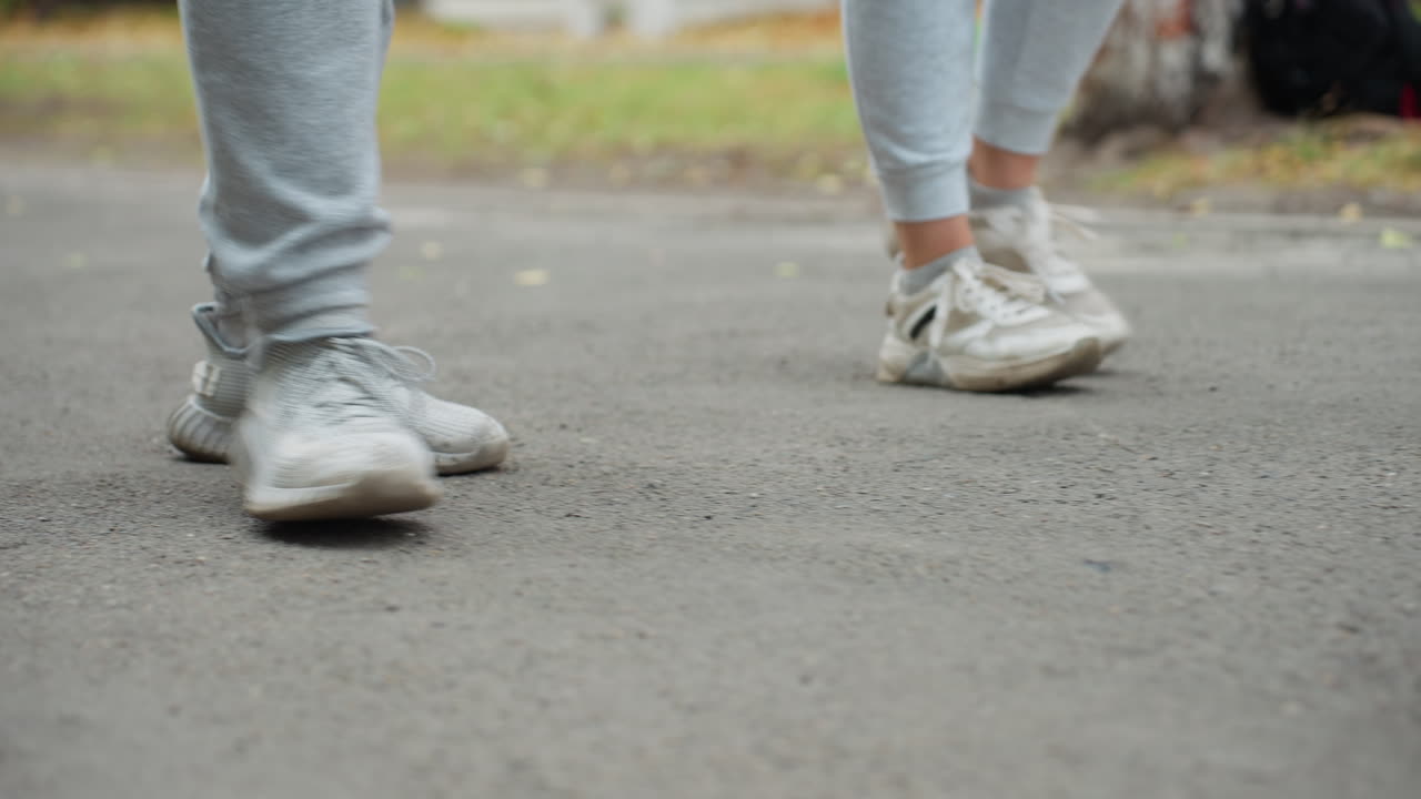 Lower angle view of two individuals walking side by side in grey joggers and sneakers, captured mid-step on paved road with greenery in blurred background