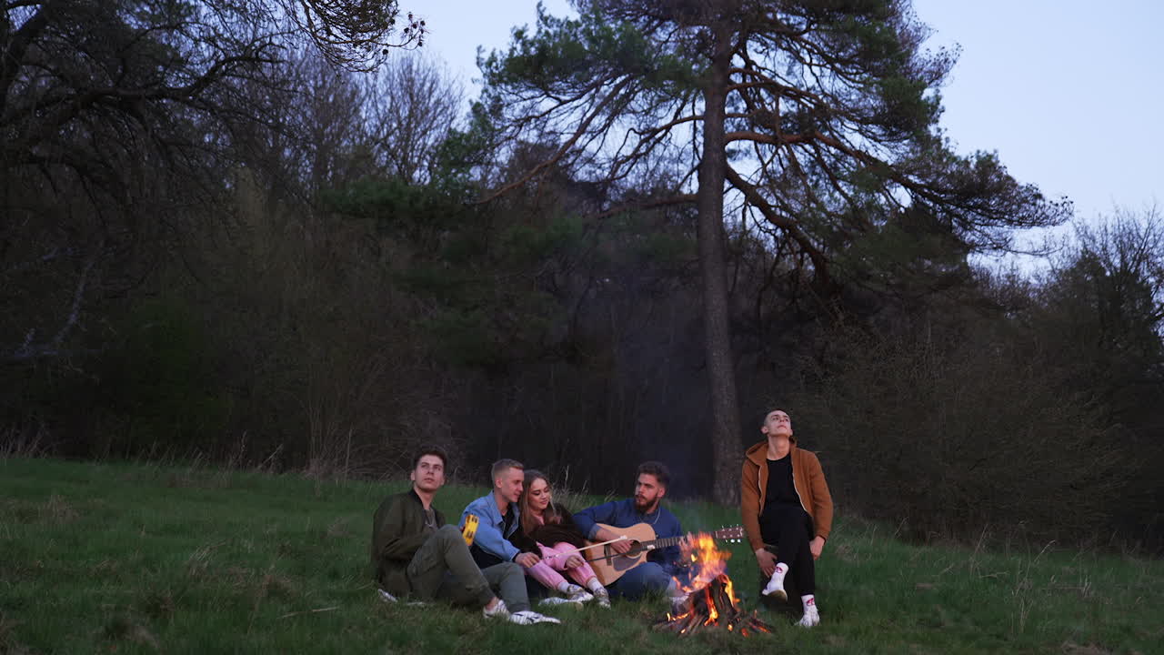 Evening picnic near the bonfire in the company of friends. Young people playing musical instruments and singing songs. Couple sitting in the middle roasting marshmallows.