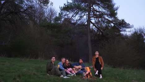 evening picnic near the bonfire in the company of friends. young people playing musical instruments and singing songs. couple sitting in the middle roasting marshmallows.