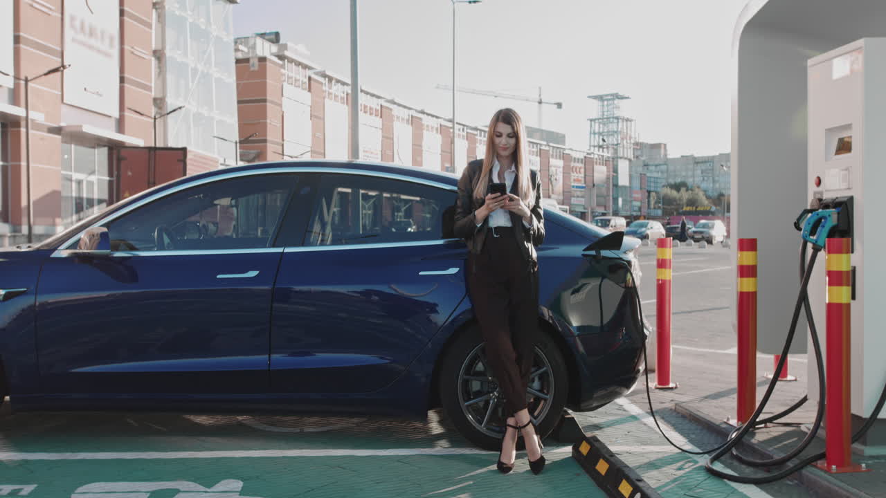 Donna in piedi con il telefono vicino alla sua auto elettrica mentre aspetta che il veicolo si carichi; donna che connette un'auto.