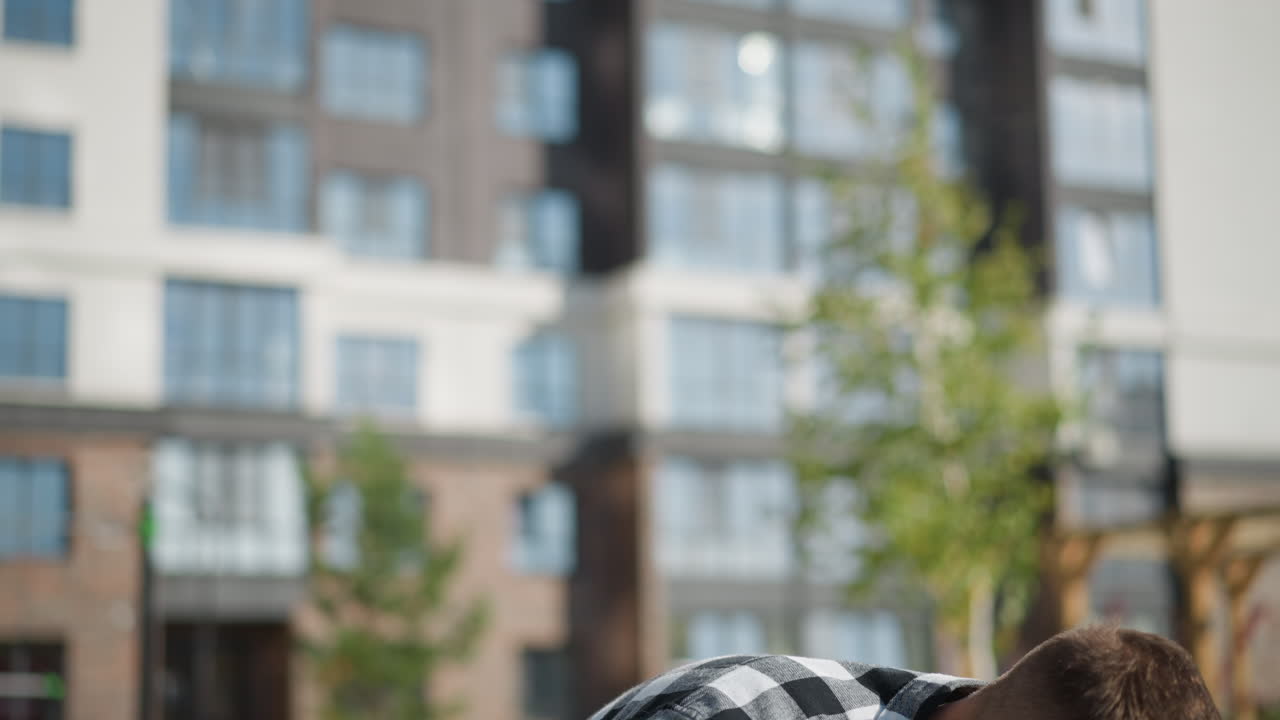 calm fair man in plaid shirt sits under bright sun reaches into front pocket for medicine with blur view of greenery and tall residential building in background