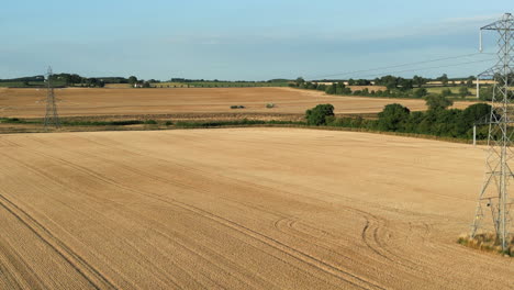 Establishing-Drone-Shot-of-Fields-of-Wheat-with-Pylons-and-Power-Lines-and-Combine-Harvester-and-Tractor-with-Trailer-in-the-Distance-at-Golden-Hour-UK