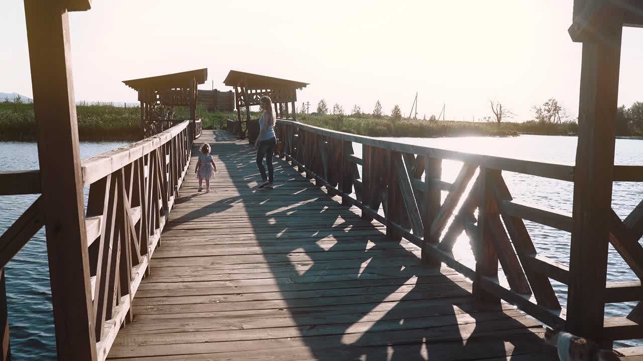 joven madre hermosa va con un niño en un puente de madera durante la puesta de sol cerca del lago