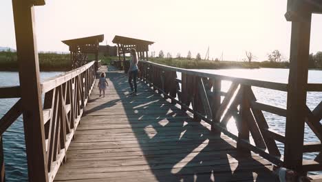 Junge-Schöne-Mutter-Geht-Mit-Einem-Kind-Bei-Sonnenuntergang-In-Der-Nähe-Des-Sees-Auf-Eine-Holzbrücke