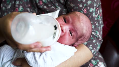 adorable close up shot of a newborn infant baby cradled in his mother’s arms feeding on a bottle of breast milk