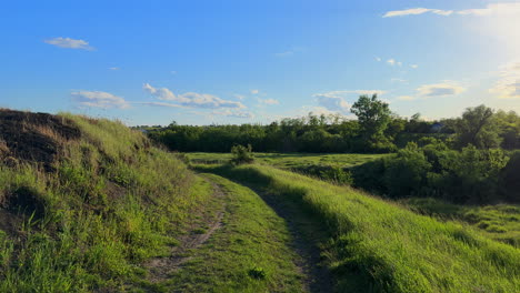 Following-Dirt-Rural-Trail-Path-Through-Grassy-Field-on-a-Sunny-Day,-Clear-Blue-Sky