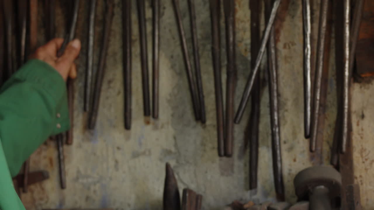 A blacksmith selects a pair of tongs from his workshop's wall, test them on a piece of metal