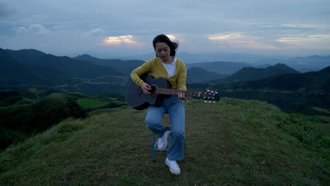 a woman strums her guitar on a hilltop, the sound of her music filling the air as the mountains provide a stunning backdrop to her performance