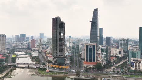drone view over central ho chi minh city, vietnam, showing bitexco financial tower and saigon one tower near the bến nghé canal and historic mong bridge, with dense cityscape beyond the waterway