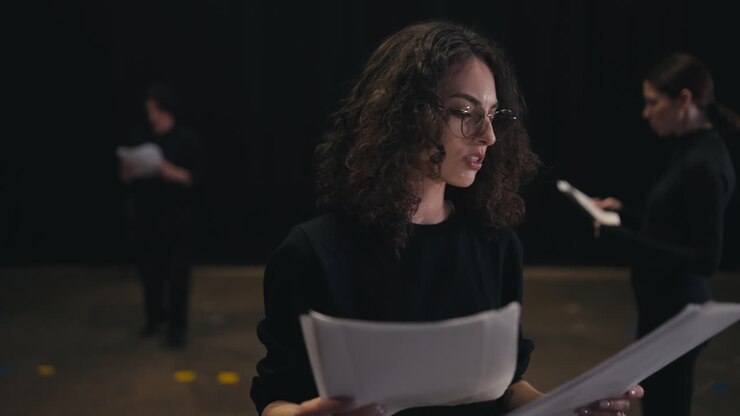 A confident girl with brown hair and round glasses in a black uniform suit reads her words and the plot of the play from sheets of paper during her preparation and rehearsal on stage in the theater