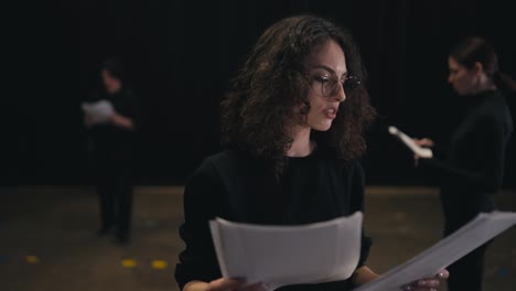 a confident girl with brown hair and round glasses in a black uniform suit reads her words and the plot of the play from sheets of paper during her preparation and rehearsal on stage in the theater