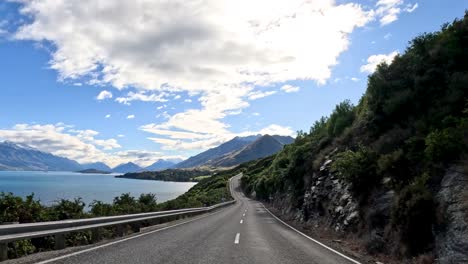 a serene drive through mountainous terrain by lake wakatipu under a bright, partly cloudy sky in queenstown, new zealand