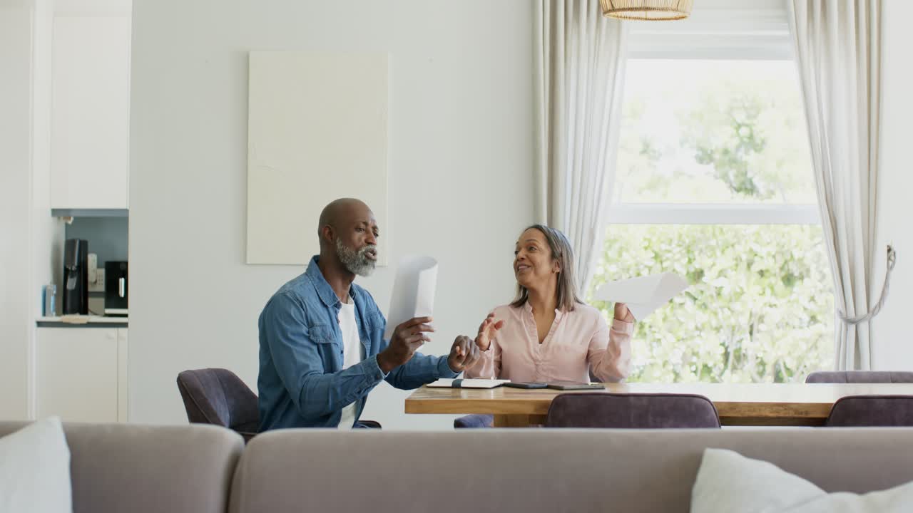 Diverse couple holding pages, reviewing documents, agreeing on details at dining table, smiling