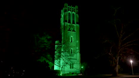 beaumont tower on the campus of michigan state university lit up at night in green in honor of the victims of the february, 2023 mass shooting with ws walking forward through trees