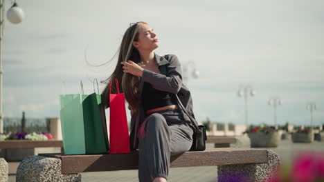 woman seated on outdoor bench with three colorful shopping bags beside her, crossing her legs and adjusting her hair while posing stylishly on a sunny day, electric poles visible in the background