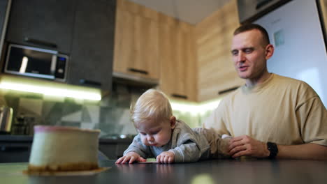 blond baby boy lies on the table trying to get to the cake in front of him. smiling dad sitting beside holds a kid.