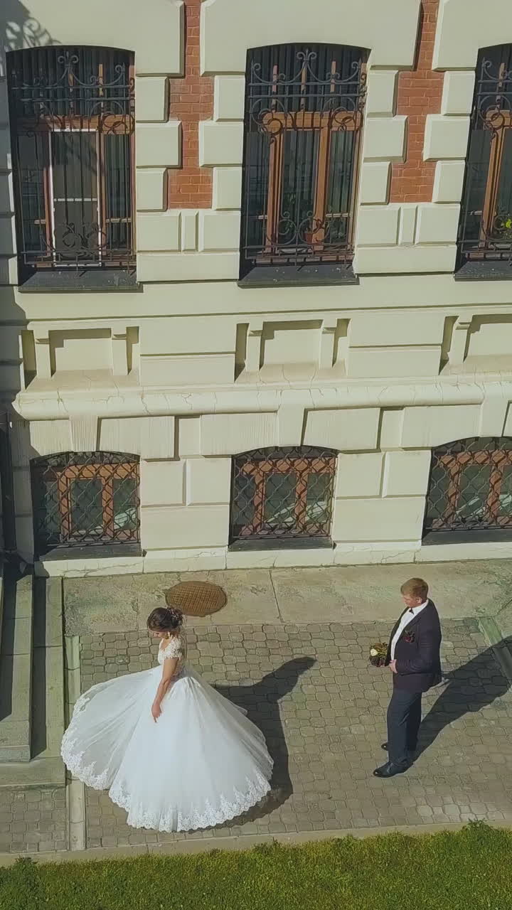 happy bride in long dress dances spinning near groom between modern building and green lawn on sunny day upper view