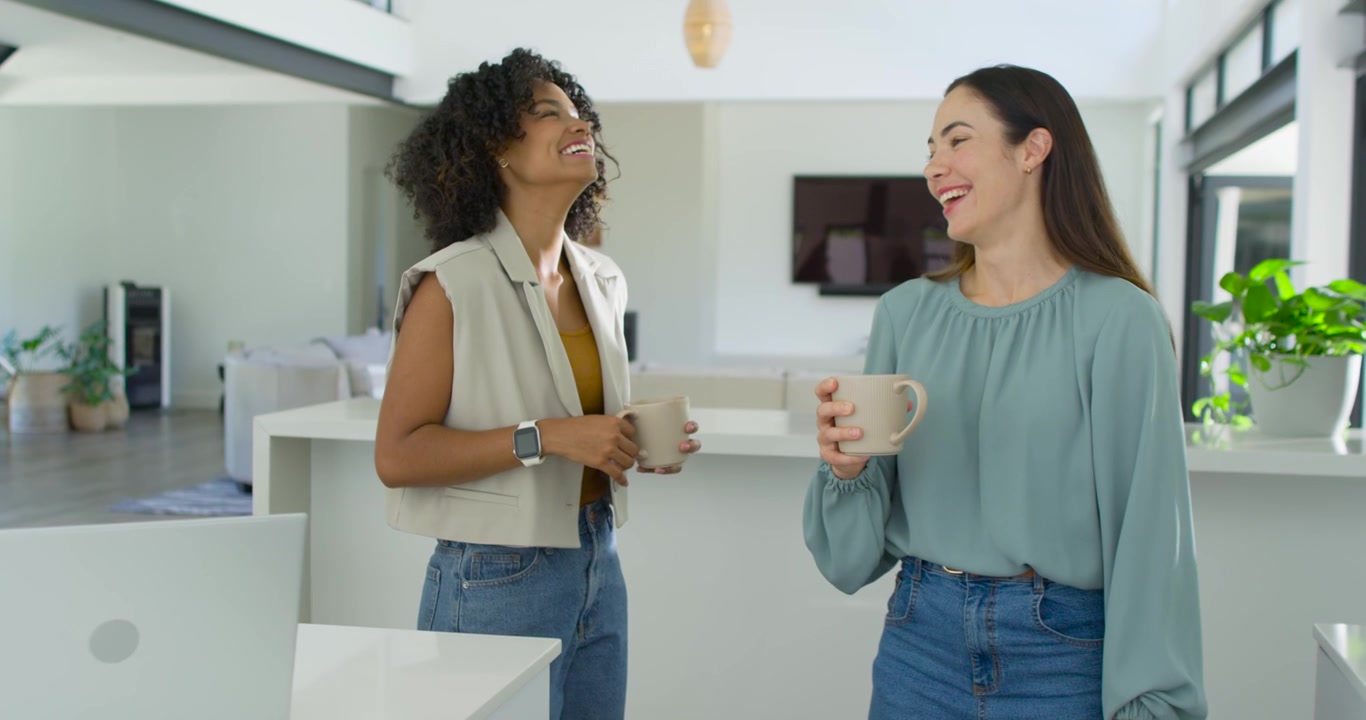 Diverse female friends, noticing laptop on island, chatting and laughing while holding ceramic mugs
