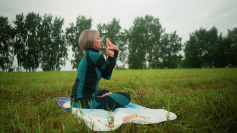 woman seated on yoga mat in misty field practicing yoga with eyes closed, hand twisting and thumb touching forehead, set under cloudy sky with trees lined up on the horizon