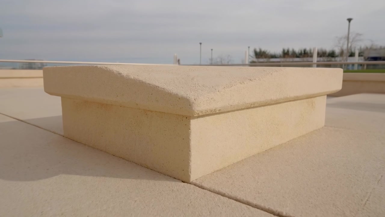 Vista de baixo ângulo, ao nível do solo, revelando uma cimalha quadrada de concreto bege repousando sobre um pedestal a condizer numa praça pública, com pavimentação texturizada, sombra suave, tons neutros e linhas geométricas limpas.