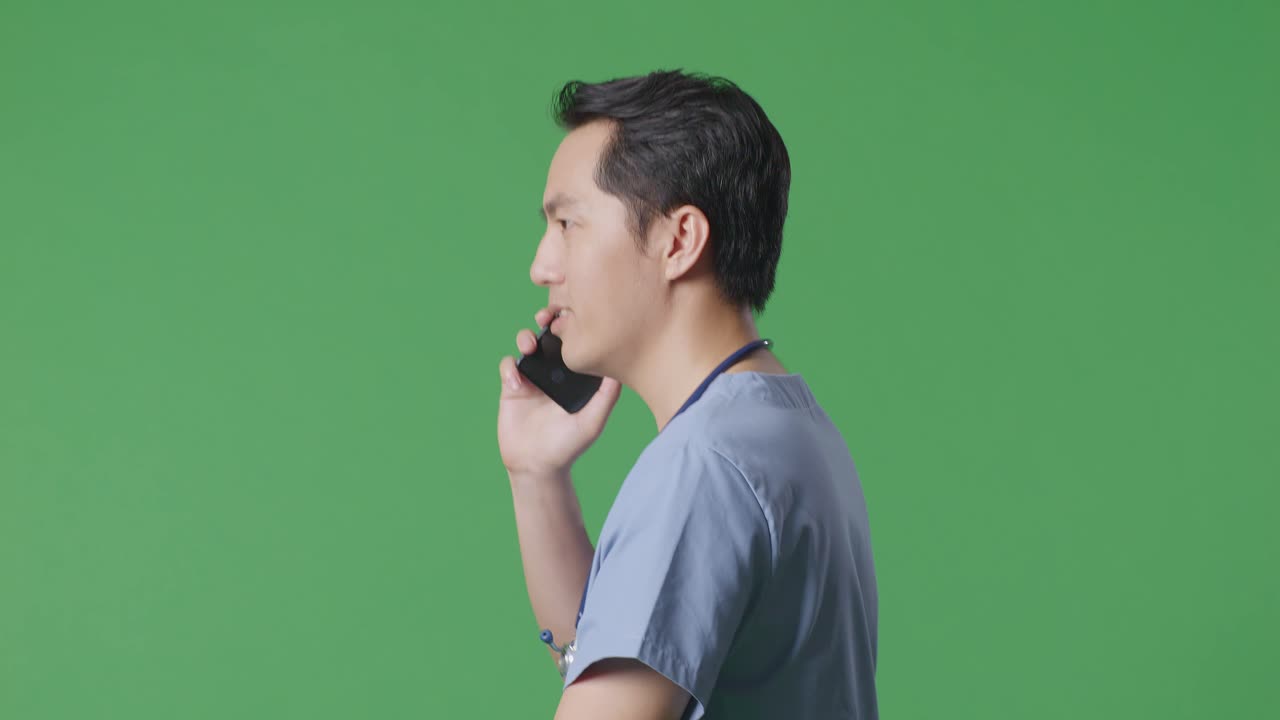 으로 가게 보이는 아시아 남성 의사가 스테토스코프를 들고 병원에서 녹색 화면 배경에서 는 동안 스마트폰으로 이야기하고 있습니다.