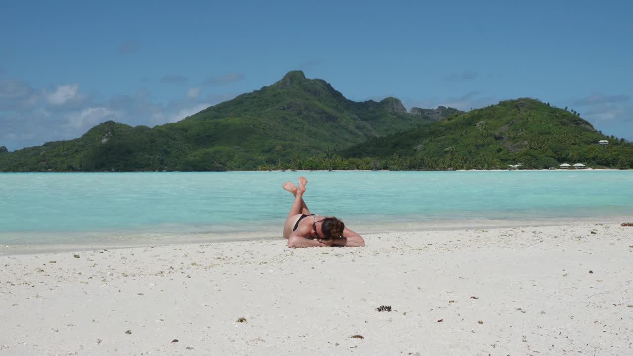 Young caucasian woman in bikini and sunglasses laying down, sunbathing and relaxing on a white sand tropical beach with feet in the water and green pacific island behind in Maupiti, French Polynesia.