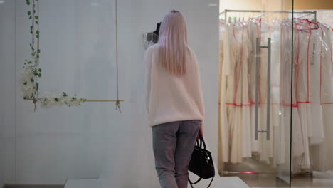 youthful lady inspecting intricate bridal gown behind glass display, soft reflections on lace bodice and tulle skirt, blurred boutique backdrop, mood of admiration and quiet anticipation