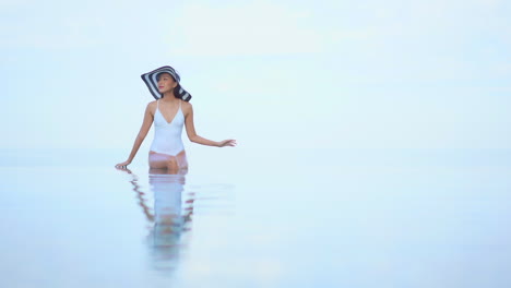 Eine-Junge-Frau-Sitzt-Mit-Blick-Auf-Die-Kamera-Am-Rand-Des-Infinity-Pools,-Rückt-Ihren-Sonnenhut-Zurecht-Und-Hat-Dabei-Die-Beine-übereinandergeschlagen