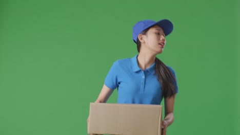 푸른 유니폼을 입은 아시아 여성 택배기사가 스튜디오에서 녹색 스크린 배경에 상자를 배달하는 동안 달리고 있습니다.
