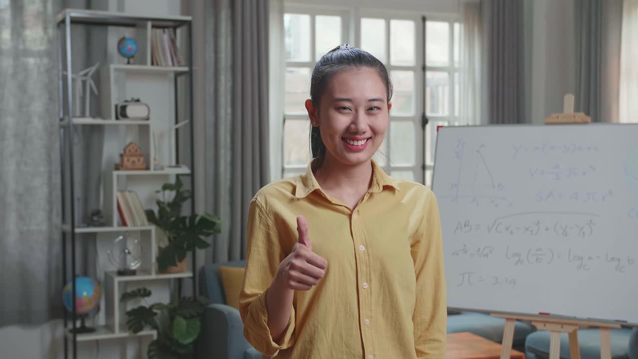 Insegnante asiatica sorridente che mostra il gesto del pollice in su alla telecamera mentre insegna matematica a casa.