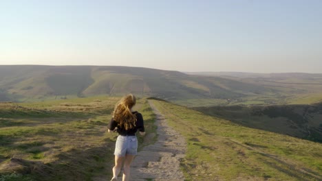 Toma-Estabilizada-De-Una-Joven-Rubia-Trotando-A-Lo-Largo-Del-Camino-En-La-Parte-Superior-De-Mam-Tor,-Castleton,-Distrito-De-Los-Picos,-Inglaterra,-Vista-De-Verdes-Colinas-Y-Cielos-Azules