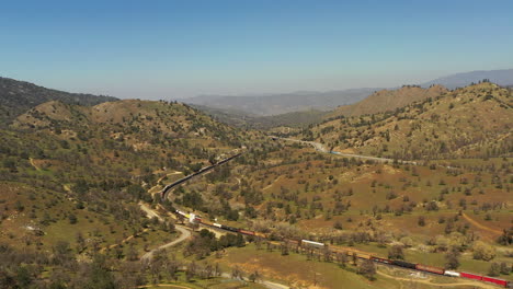 a long serpentine train winds through the tehachapi foothills below the highway traffic - aerial view