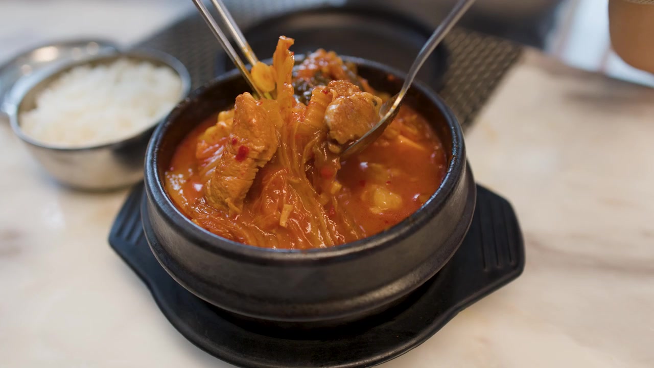 Sumpit logam mengambil daging babi dan kimchi dari mangkuk sup tahu pedas merah yang mengepul.