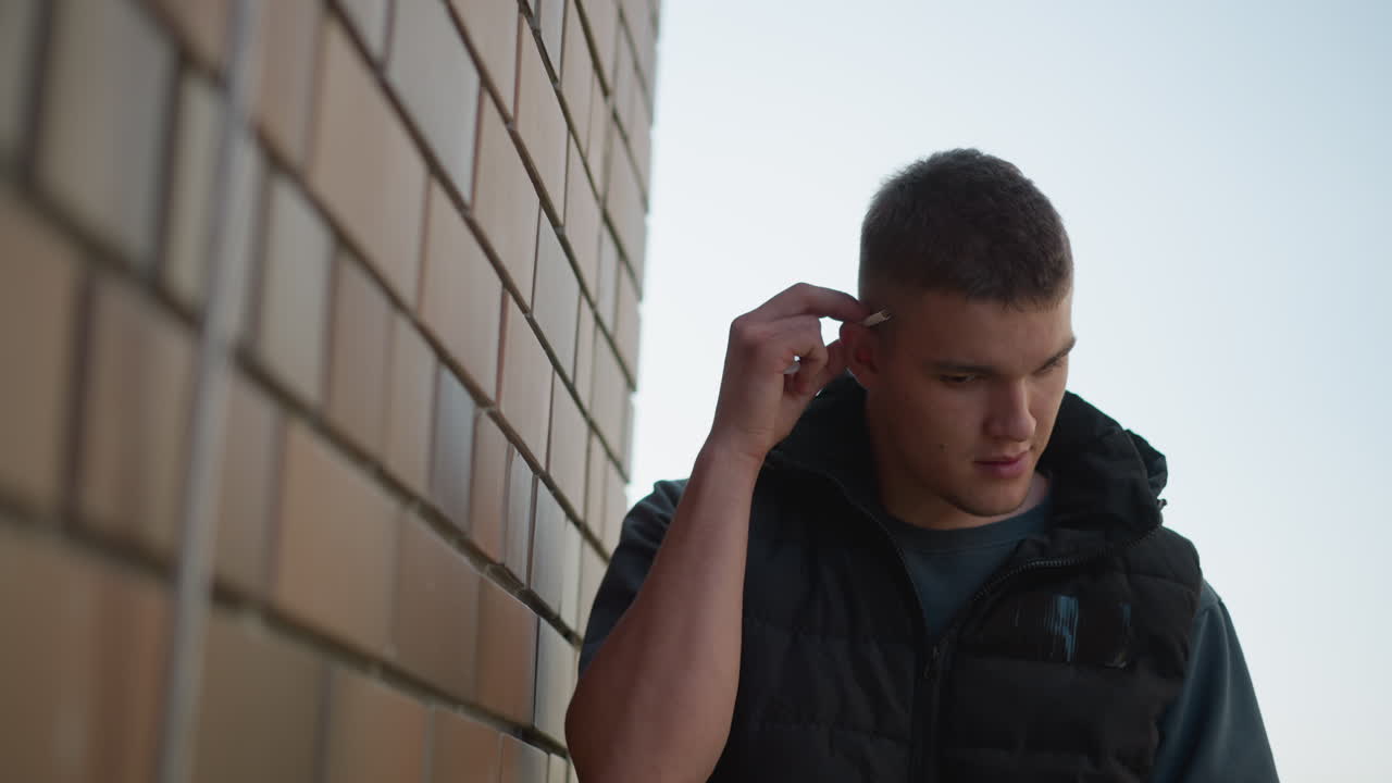 man stands beside brick wall reaching to take cigarette from ear with intention to light and smoke, dressed in black padded vest and dark shirt under bright outdoor light with blur background