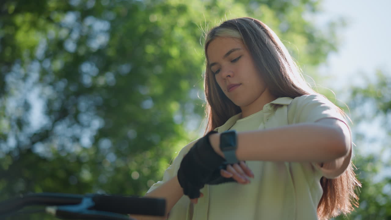 Young adult adjusting black biker glove on right hand with focused expression, standing beside bicycle as sunlight filters through green leaves