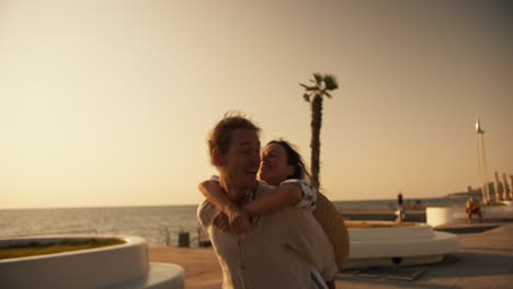 A-happy-girl-in-a-white-and-blue-shirt-approaches-her-boyfriend-from-behind-and-he-lifts-her-into-his-arms-on-a-beach-with-palm-trees-near-the-sea-at-dawn-in-summer.-Happy-couple-guy-and-girl-fooling-around-on-the-beach-near-the-sea