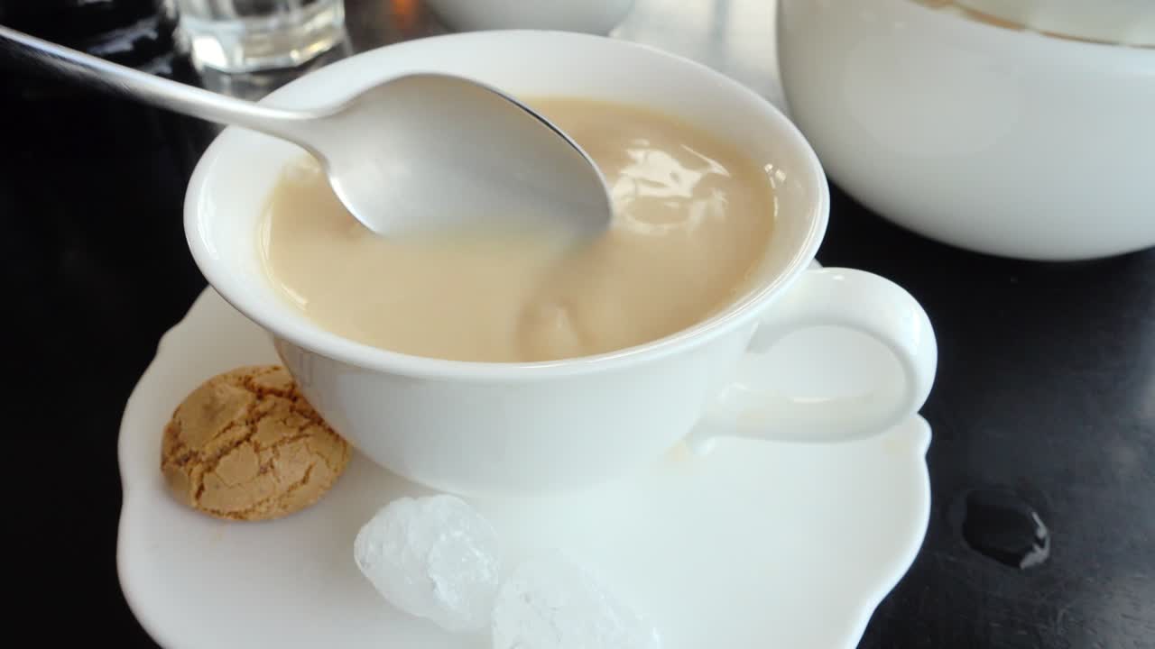 Stirring a hot cup of tea with milk to cool it down a bit. A few teadrops on saucer and table.