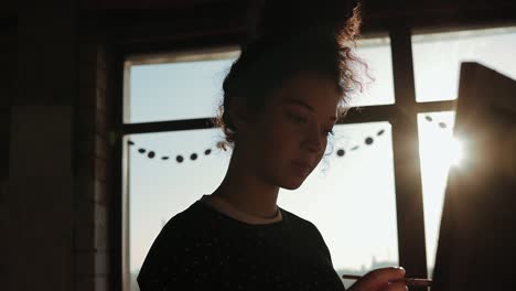 Shaded-by-the-sun-behind-young-talented-female-artist-drawing-on-easel-in-an-art-studio.